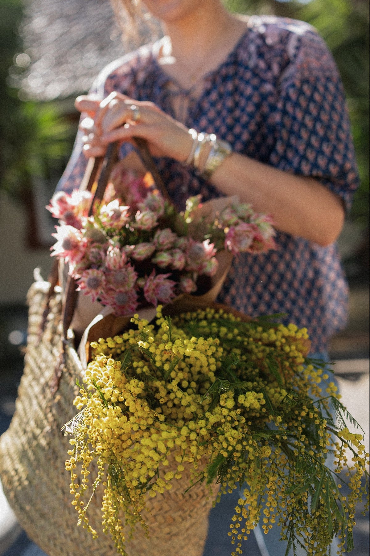 Woman wearing a navy hand block printed blouse, carrying woven baskets filled with pink and yellow flowers.