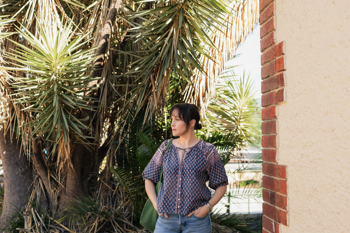 Woman wearing the Lisette navy hand block printed blouse with jeans and green crochet tote bag in front of palms. 
