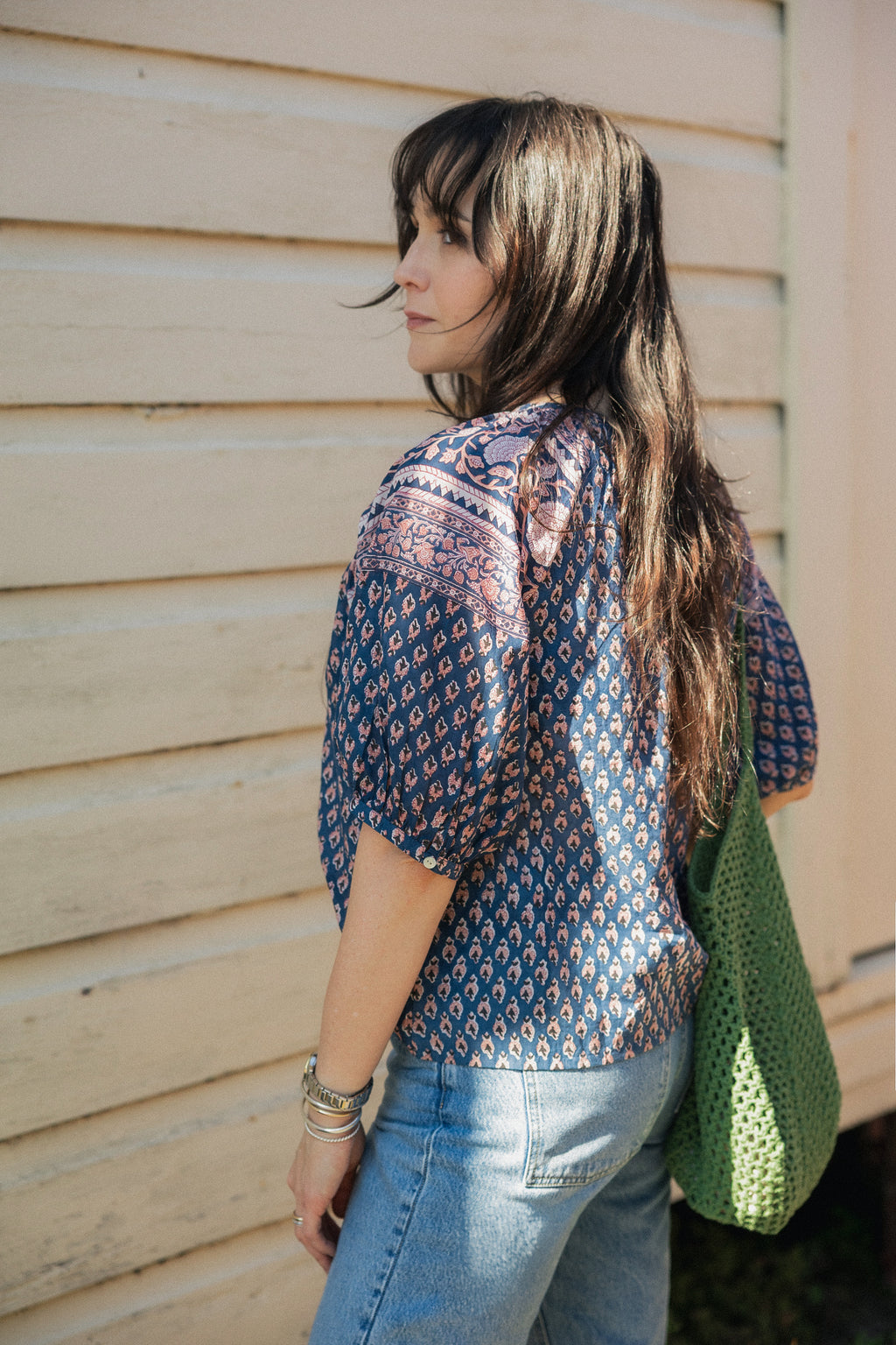 Woman standing in sun wearing the Lisette navy hand block printed blouse with jeans and green crochet tote bag.
